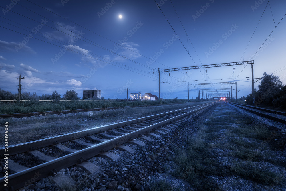 Railroad and blue sky with moon and clouds at night. Summer rural industrial landscape with railway station, trees, grass at twilight. Railway platform at dusk. Freight transportation. Heavy industry