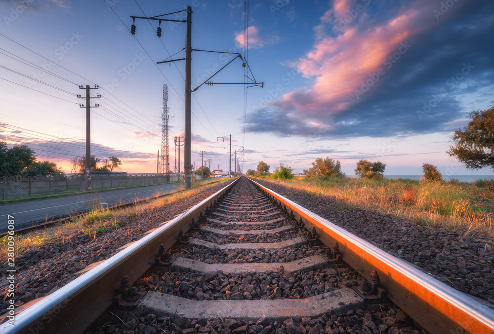 Railroad and beautiful sky at sunset in summer. Rural industrial landscape with railway station, blue sky with colorful clouds and orange sunlight, road.  Railway platform. Sleepers. Heavy industry.