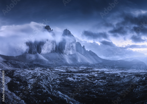 Mountains in fog at beautiful night. Dreamy landscape with mountain peaks, stones, grass, purple sky with blurred low clouds, stars and moon. Rocks at dusk. Tre Cime in Dolomites, Italy. Italian alps © den-belitsky