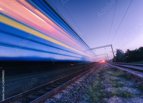 High speed passenger train in motion on the railroad at summer night. Moving blurred modern commuter train at dusk. Industrial landscape with railway station and purple sky. Transport. Travel