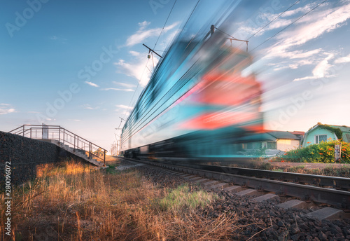 High speed passenger train in motion on the railroad at summer evening. Moving blurred modern commuter train at sunset. Industrial landscape with railway station and blue sky. Transport. Travel