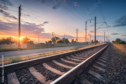 Railroad and beautiful sky at sunset in summer. Rural industrial landscape with railway station, blue sky with colorful clouds and orange sunlight, road. Railway platform. Sleepers. Heavy industry.