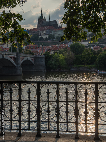 Prague castle on summer evening. Back lit seen across Vltava river with a brodge, cast iron railing and sunlit green eaves in foreground photo