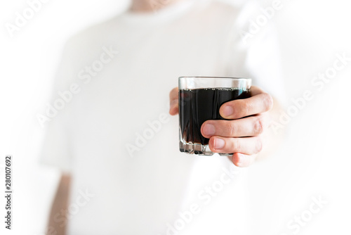 Man gives a glass of cola to camera isolated on a white background. Dark fizzy soda. Soda black in male's hand in white t-shirt