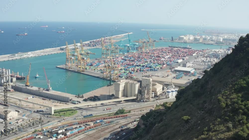 Aerial view of Barcelona industrial harbour with cranes and containers, aside mountain and automobiles moving highway. Drone shot of large cargo port in Barcelona on background of scenic seascape Aerial view of Barcelona industrial harbour with cranes and containers, aside mountain and automobiles moving highway. Drone shot of large cargo port in Barcelona on background of scenic seascape
