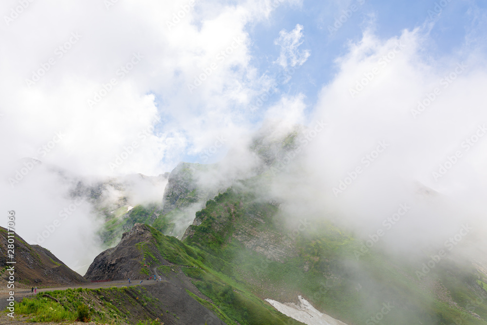 Mountain landscape. High mountains with green grass, the road through the mountains. White clouds around the mountains, blue sky behind them. Horizontal image, day light. 