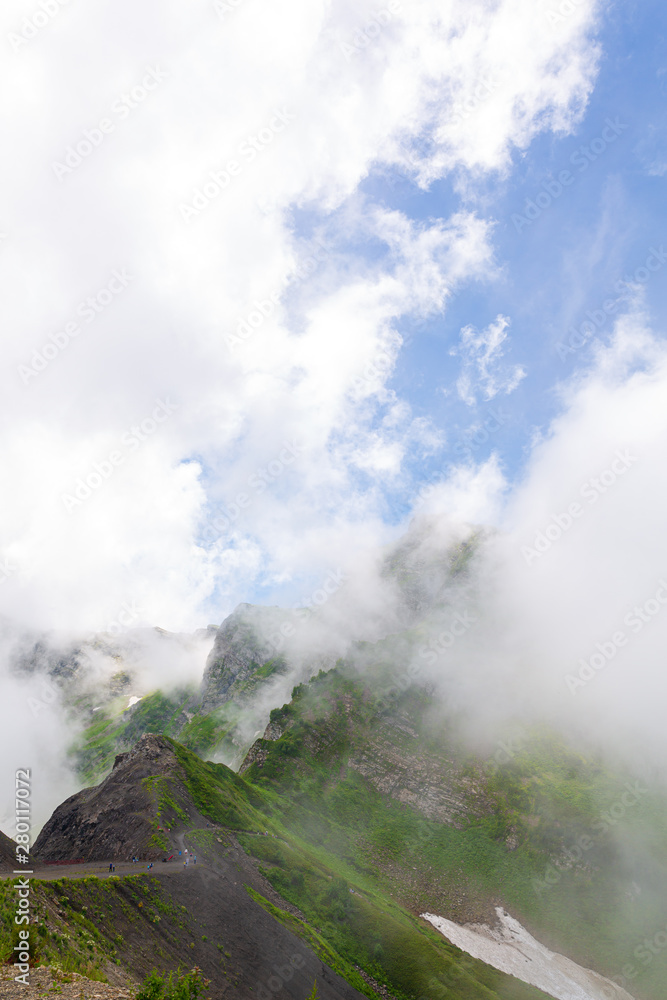 Mountain landscape. High mountains with green grass, the road through the mountains. White clouds around the mountains, blue sky behind them. Vertical image, day light. 