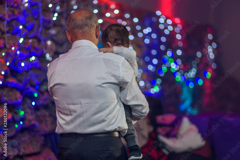 Back view happy grandfather holding her baby grandson in arms. Old senior dad with little son on a disco lights background. Back view happy grandfather holding her baby grandson in arms. Old senior dad with little son on a disco lights background.