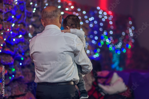 Back view happy grandfather holding her baby grandson in arms. Old senior dad with little son on a disco lights background.