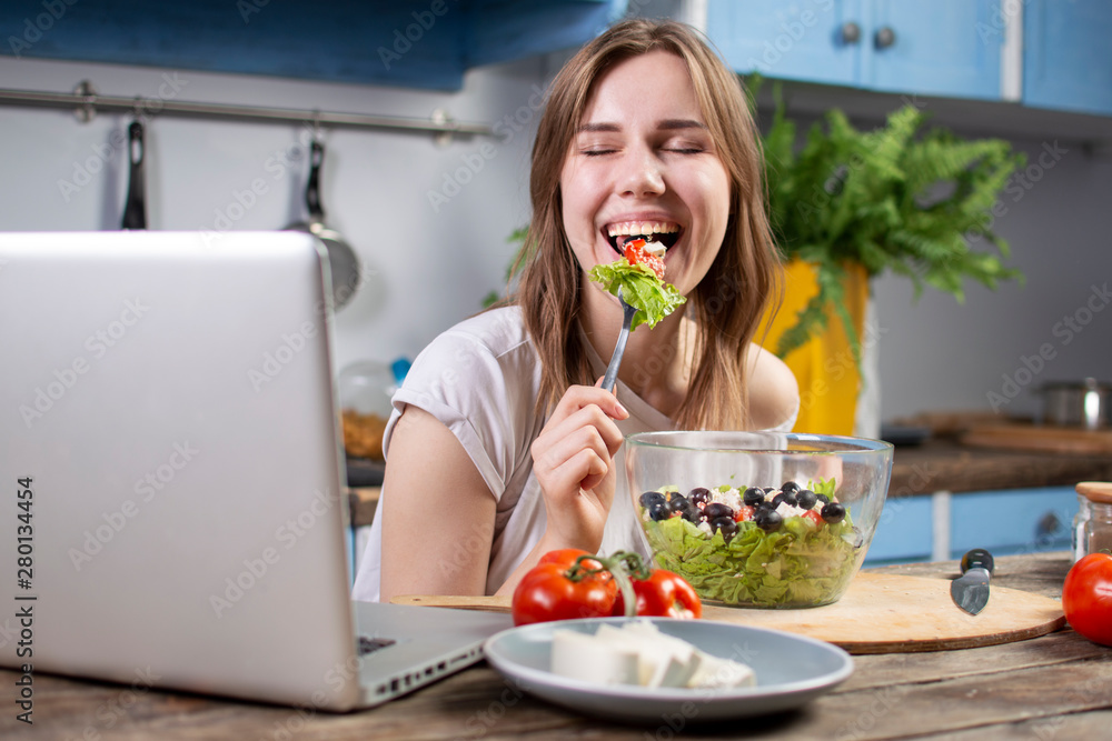 Young girl eating a vegetarian salad at home in the kitchen and watching at a laptop, she eats healthy food at a wooden table Young girl eating a vegetarian salad at home in the kitchen and watching at a laptop, she eats healthy food at a wooden table
