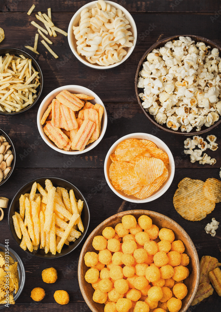 All classic potato snacks with peanuts, popcorn and onion rings and salted pretzels in bowl plates on wooden background. Twirls with sticks and potato chips and crisps with nachos and cheese balls.