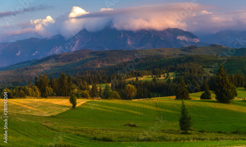 Fototapeta Naklejka Na Ścianę i Meble -   Tatra Mountains. Mountain peaks towering over green grassy hills. Above the peaks, beautiful storm clouds illuminated by the setting sun