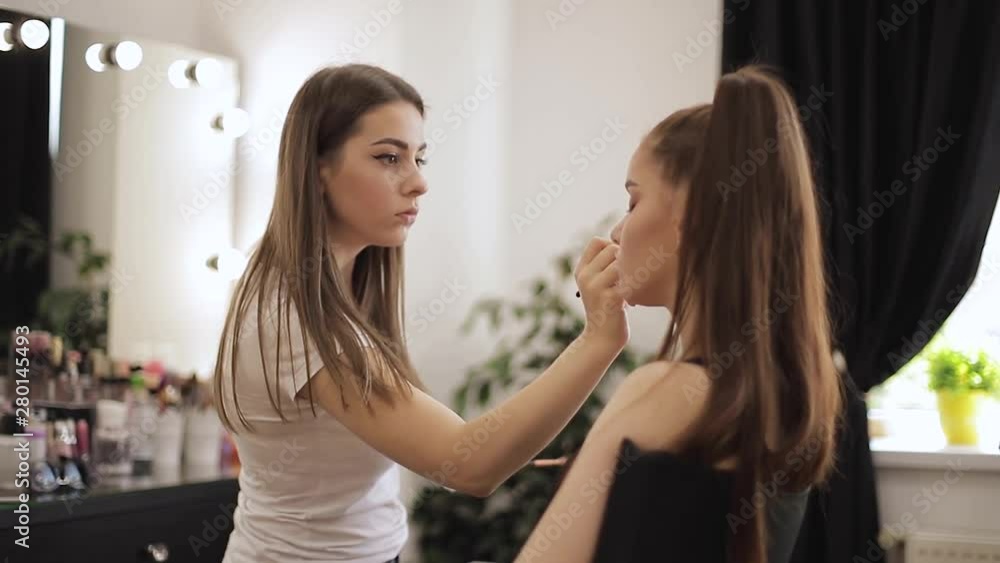 Beautiful femalemakeup artist doing makeup for a young redhead girl in a beauty salon sitting in front of a large mirror. Concept of preparation for the holiday and meeting Beautiful femalemakeup artist doing makeup for a young redhead girl in a beauty salon sitting in front of a large mirror. Concept of preparation for the holiday and meeting