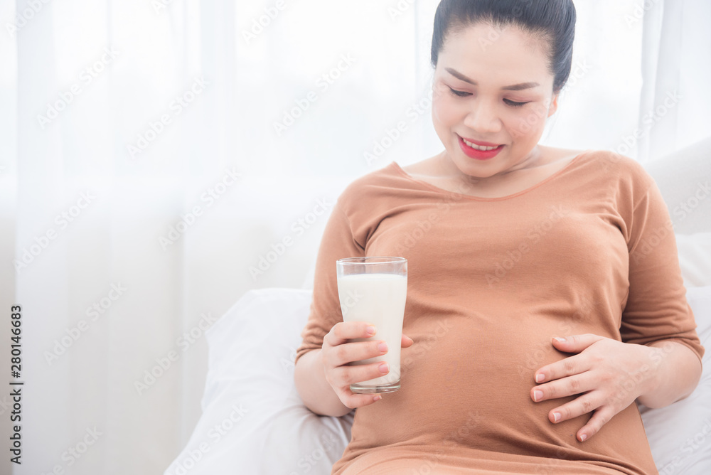 Pregnant woman sitting on bed ,holding a glass of milk and smiles.Focus at milk glass and pregnant belly. Pregnant woman sitting on bed ,holding a glass of milk and smiles.Focus at milk glass and pregnant belly.