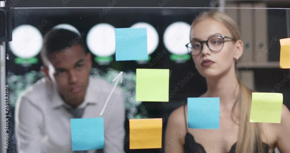 Young woman with glasses in a modern office, draws arrows infographic on the glass board with sticky notes, african man look at the board. Young woman with glasses in a modern office, draws arrows infographic on the glass board with sticky notes, african man look at the board.