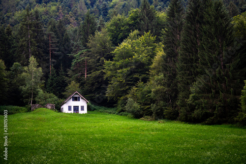 Mountain landscape with white barn or cabin in the distance across a green meadow. 