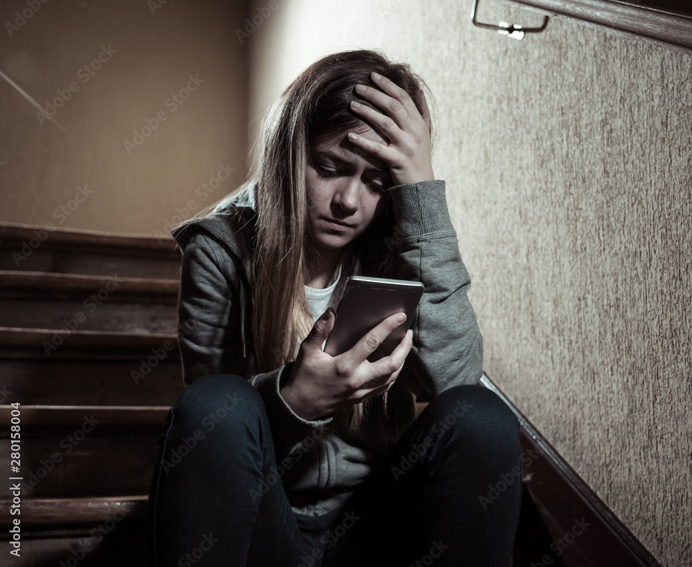 Sad depressed unhappy teenager girl suffering from cyberbullying by mobile smart phone sitting alone.