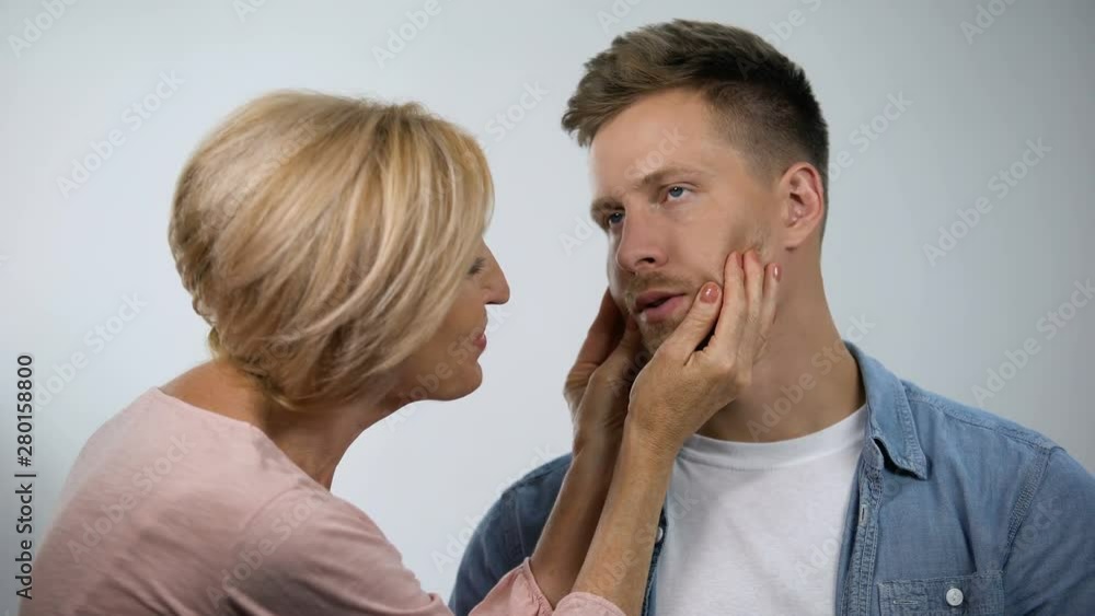 Smiling mother pinching irritated son cheeks, adult child care, overprotection Smiling mother pinching irritated son cheeks, adult child care, overprotection