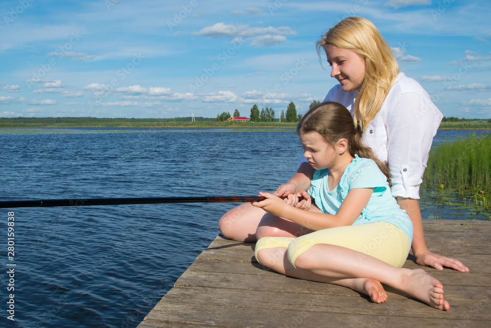 Fototapeta premium mom and daughter on the pier, against the backdrop of a beautiful landscape, hold a fishing pole to catch fish, close-up, there is a place for an inscription
