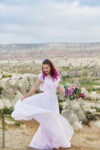 Woman with a beautiful bouquet of flowers in her hands dance on the mountain in the rays of the dawn sunset. Beautiful white long dress on the girl body. Perfect bride with pink hair dance