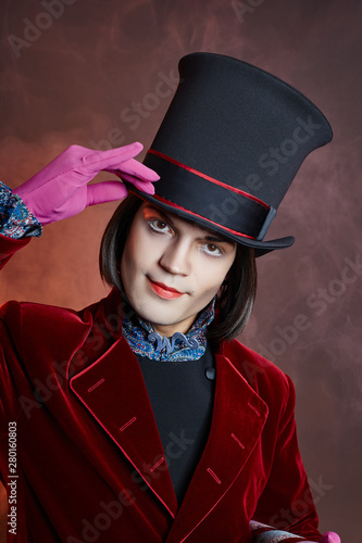 Fabulous circus man in a hat and a red suit posing in the smoke on a colored dark background. A clown at a party, man gentleman from a fairy tale. Wizard with a cane, ice cream and chocolate in hands