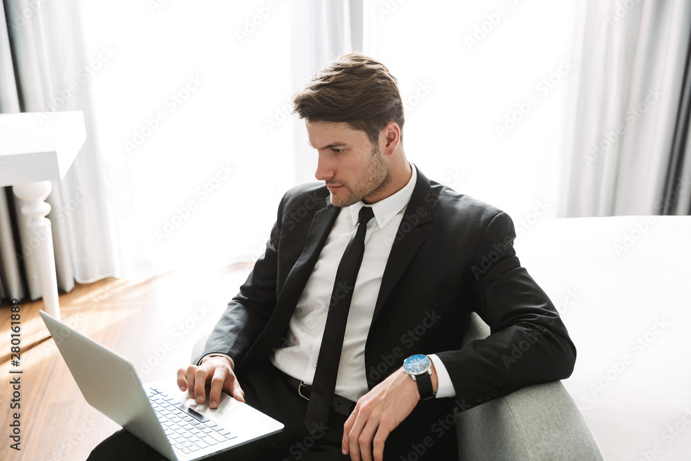 Image of concentrated young man sitting on sofa with laptop computer in hotel apartment during business trip Image of concentrated young man sitting on sofa with laptop computer in hotel apartment during business trip