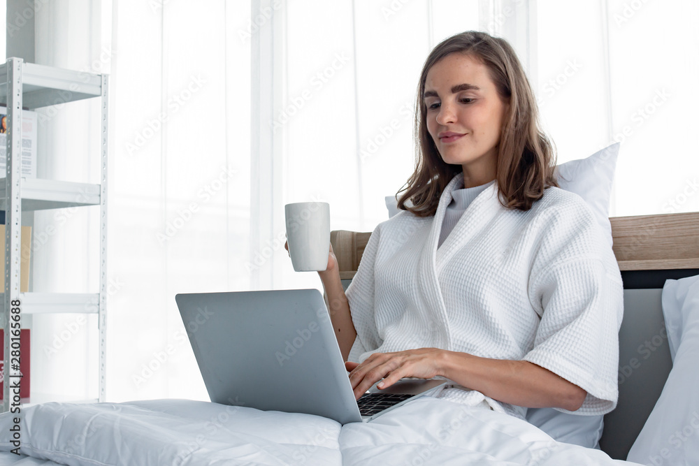 Young caucasian woman in white pajamas with white holding a cup of coffee and drink and find information from the laptop on a bed. Young caucasian woman in white pajamas with white holding a cup of coffee and drink and find information from the laptop on a bed.