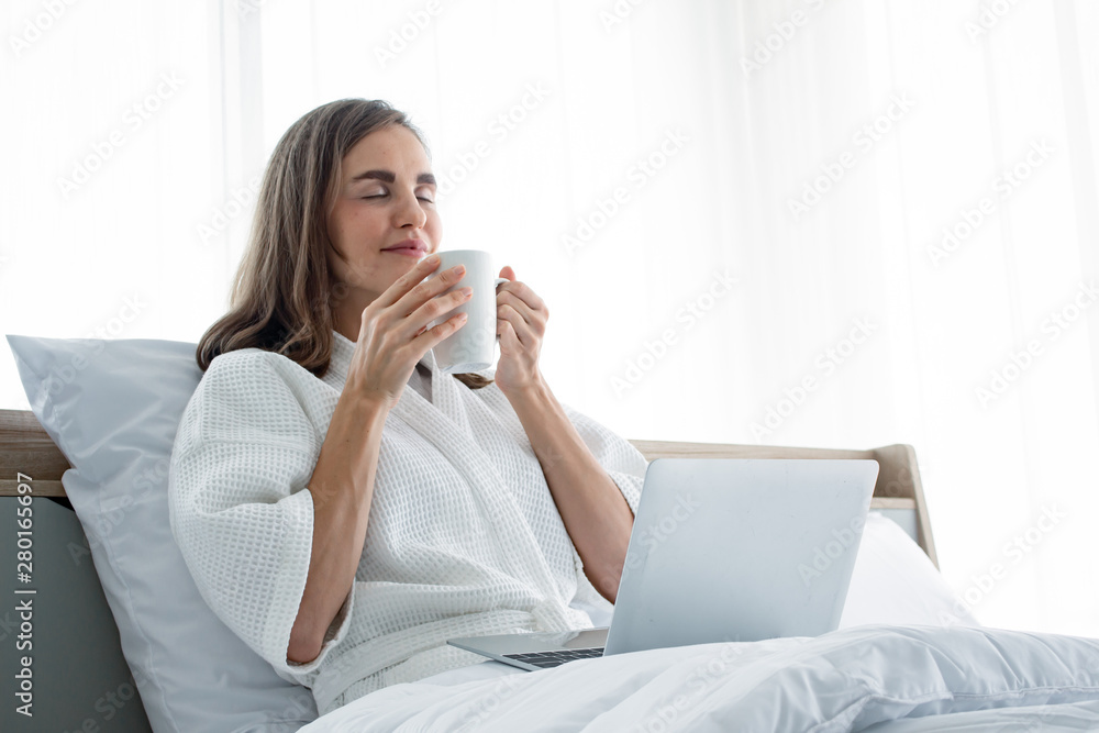 Young caucasian woman in white pajamas with white holding a cup of coffee and drink and find information from the laptop on a bed. Young caucasian woman in white pajamas with white holding a cup of coffee and drink and find information from the laptop on a bed.