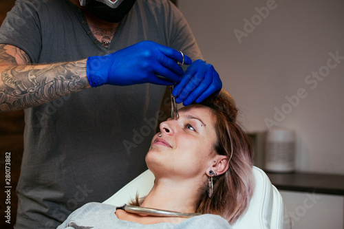 Young Woman getting pierced between her eyes. Man showing a process of piercing with steril medical equipment and latex gloves. Body Piercing Procedure photo