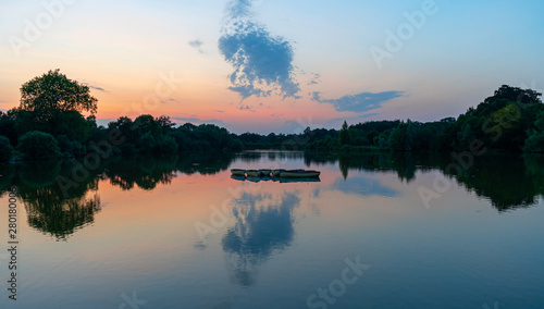 Sunset, twilight view of orange, pink and blue sky reflected in tranquill lake with rowing boats silhouette to centre of frame