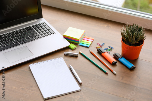 An open notebook for notes, pencils, markers, a pen, a pot with cactus, a notebook and paper clips on a wooden background (office desk).