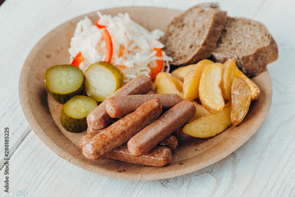 Tasty sausages with fried potatoes, cucumber, cabbage and chlobe in a paper plate on a wooden table. Tasty sausages with fried potatoes, cucumber, cabbage and chlobe in a paper plate on a wooden table.