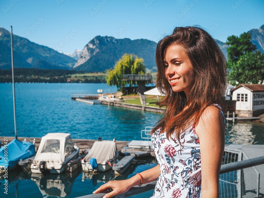 Brunette girl with long hair in a blue dress stands on the waterfront of the city of Thun, lake Geneva, transparent turquoise