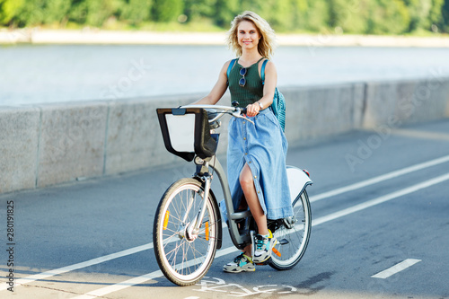 Image of young blonde in long denim skirt sitting on bike on road in city