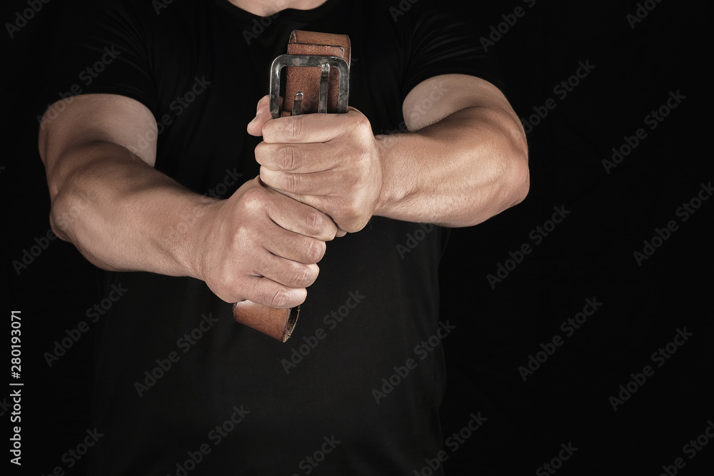 adult man in black clothes holding a brown leather belt with an iron buckle adult man in black clothes holding a brown leather belt with an iron buckle