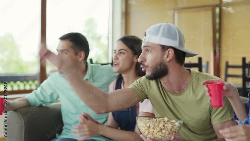 Side view of four young friends sitting on sofa at home, watching tv together and cheering during sports game. Fans eating popcorn and drinking beer. Man screaming emotionally supporting team Side view of four young friends sitting on sofa at home, watching tv together and cheering during sports game. Fans eating popcorn and drinking beer. Man screaming emotionally supporting team