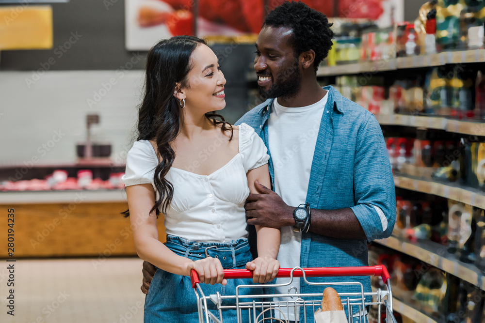 happy asian woman and handsome african american man looking at each other in supermarket