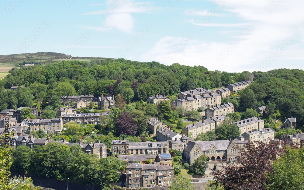 a scenic aerial view of the town of hebden bridge in west yorkshire with hillside streets of stone houses and roads between trees and a blue summer sky