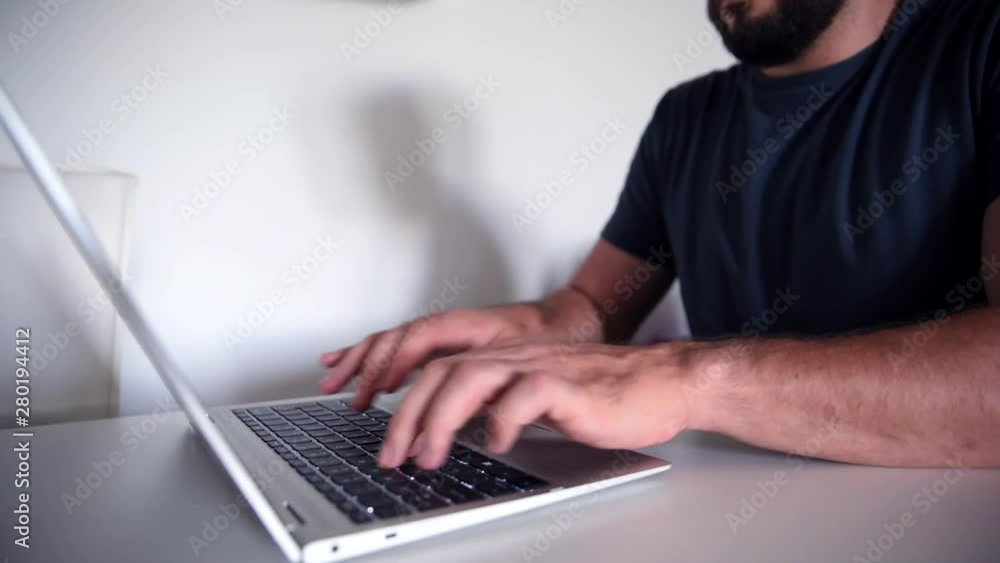 Bearded man typing rapidly on the brand new modern silver laptop placed on white table during morning captured with dolly movement Bearded man typing rapidly on the brand new modern silver laptop placed on white table during morning captured with dolly movement