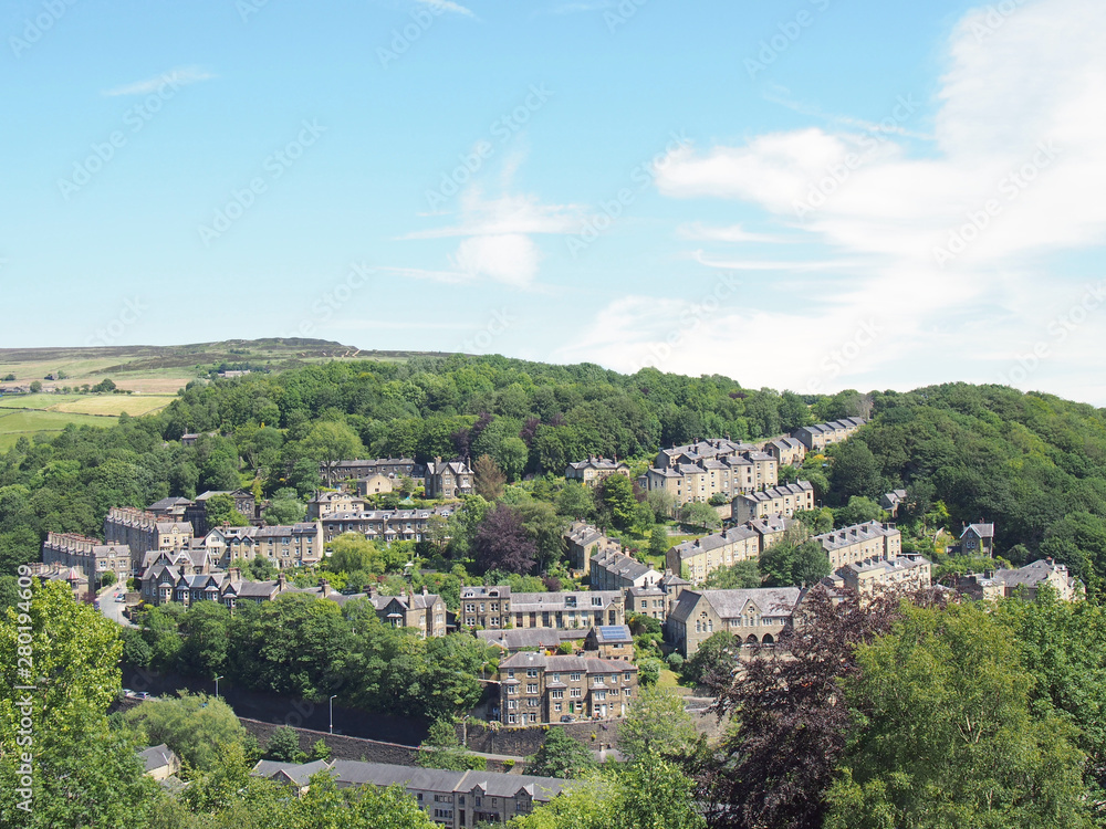 a scenic aerial view of the town of hebden bridge in west yorkshire with streets of stone houses and roads between woodland trees and a blue summer sky