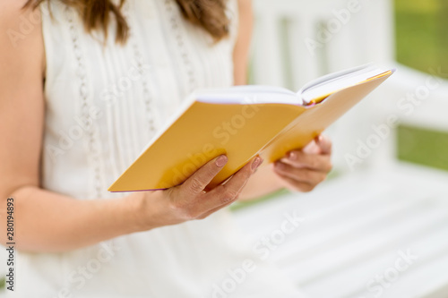 leisure, literature and people concept - close up of young woman in white dress reading book sitting on bench at summer park