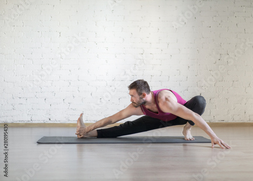 Caucasian man doing stretching, warm-up, yoga on grey mat in fitness studio, loft style, selective focus.