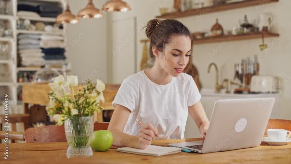 Attractive young brunette woman making some notes in the notebook from laptop computer sitting at the dining room Attractive young brunette woman making some notes in the notebook from laptop computer sitting at the dining room