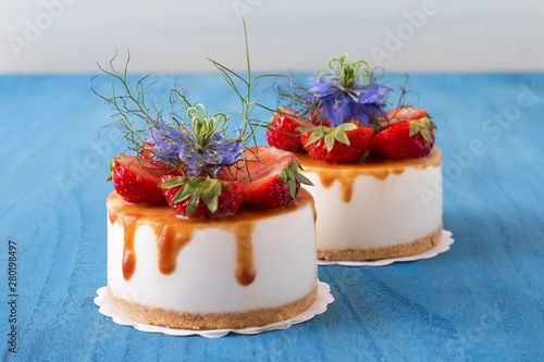 Two round no bake strawberry mini cheesecakes with salt caramel and halfs of strawberries, embellished with real blue blossoms..Blue wooden table.