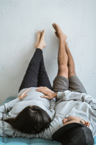 asian couple relaxing on the couch with hands hold of belly wife pregnant and feet placed on the wall
