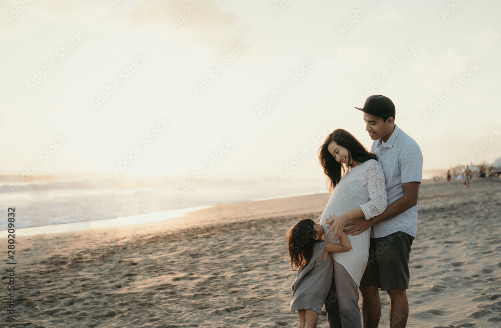 Family vacation on the beach. Father, pregnant mother, and daughter standing on the beach enjoying the sunrise Family vacation on the beach. Father, pregnant mother, and daughter standing on the beach enjoying the sunrise