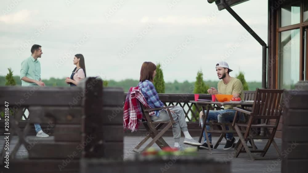 Two young couples talking on country house terrace after dinner. Emotional man telling story to girlfriend sitting at table. Cheerful couple talking over beverages leaning on railing in background Two young couples talking on country house terrace after dinner. Emotional man telling story to girlfriend sitting at table. Cheerful couple talking over beverages leaning on railing in background