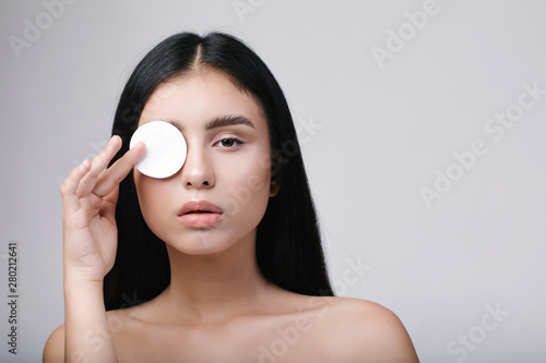 Perfect girl with nude make up and naked shoulders posing at white background with cleaning sponge, skin care concept, close up portrait. Portrait of young woman holding cotton pad near her eyes.