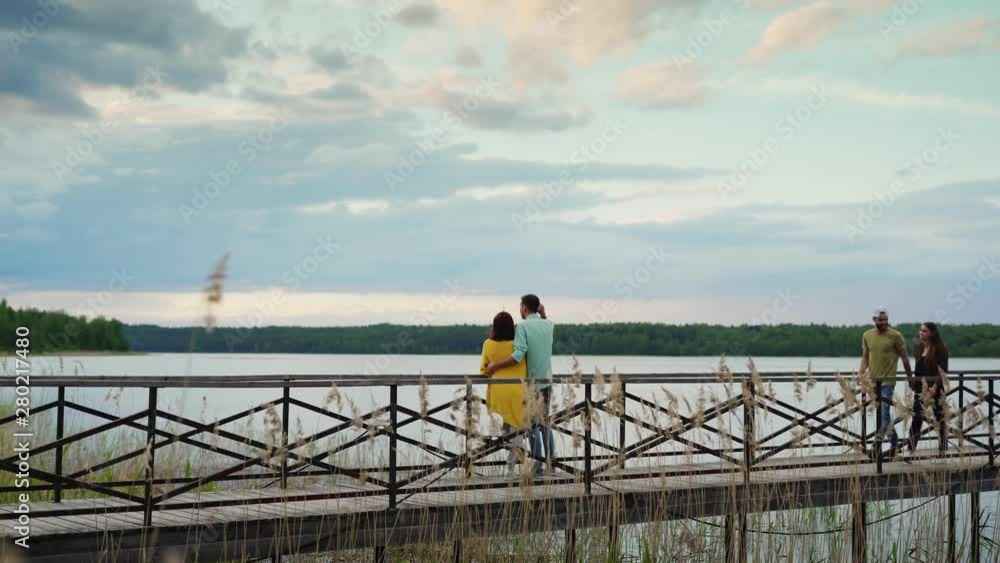 Back view of romantic young couple standing on wooden bridge and admiring beautiful river and sky view. Another couple walking by them holding hands, talking joyfully and laughing Back view of romantic young couple standing on wooden bridge and admiring beautiful river and sky view. Another couple walking by them holding hands, talking joyfully and laughing