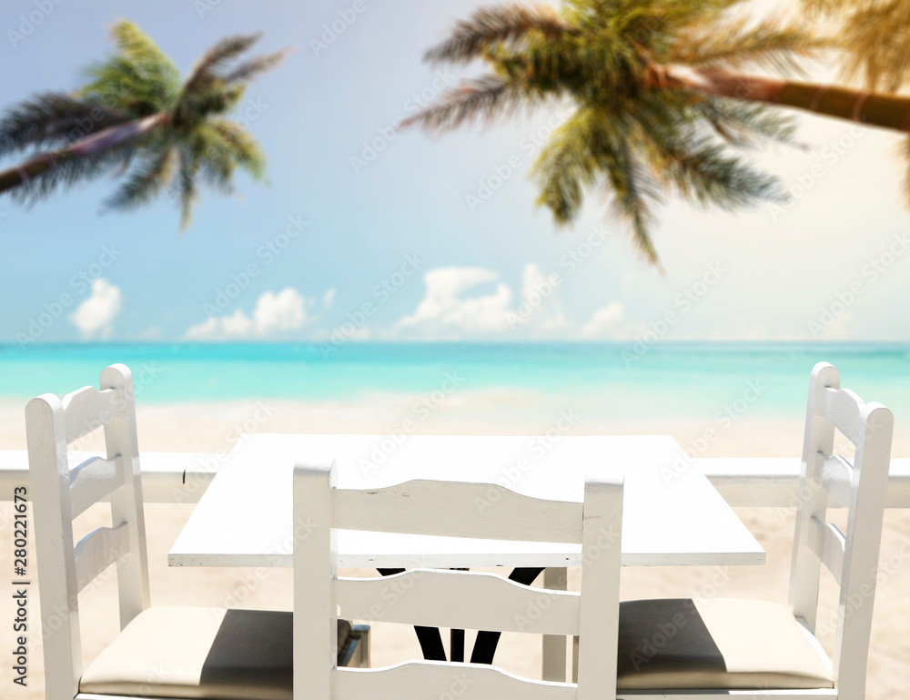 Table background with white wooden top and chairs on a sunny beach and blue ocean and sky view.